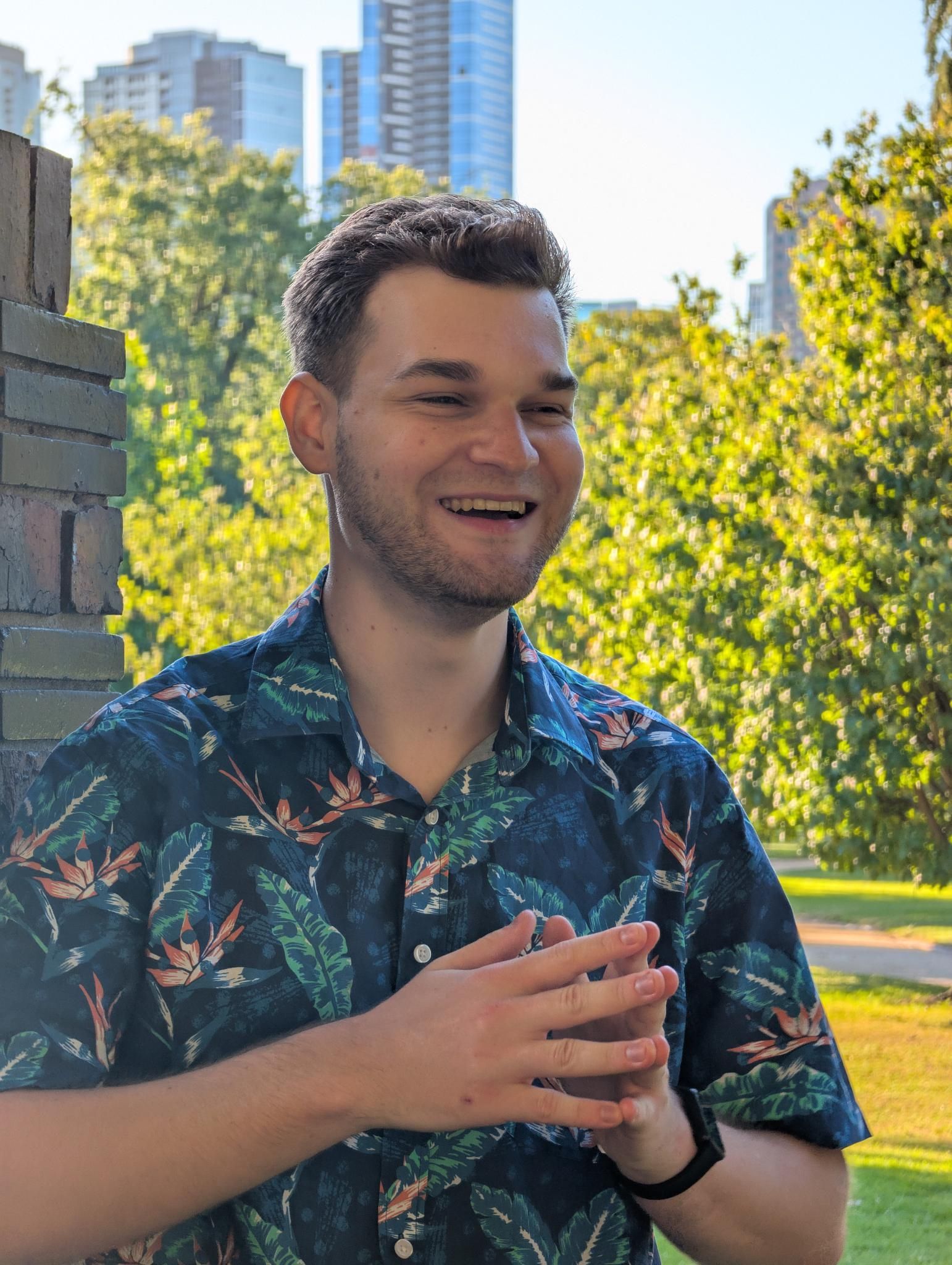 Portrait of Zach Jensz smiling, with trees and Melbourne skyscrapers in the background
